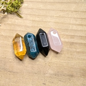 May include: Four crystal pendants in shades of yellow, teal, black, and pink. Each crystal is a different colour and has a hole at the top. The crystals are arranged on a light brown wooden surface.