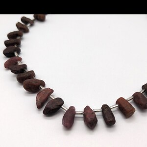 May include: A strand of dark red gemstone beads strung on a silver wire. The beads are irregularly shaped and have a rough, natural texture.