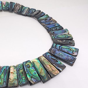 Könnte beinhalten: Nahaufnahme einer Halskette aus rechteckigen Stücken schillernder Abalone-Muscheln. Die Stücke zeigen Farbtöne von Blau, Grün und Lila. Die Halskette ist in einer gebogenen Form angeordnet, vor weißem Hintergrund.