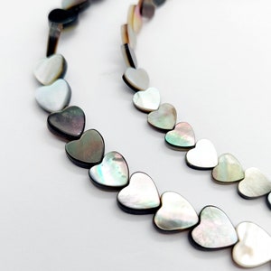 May include: Two strands of heart-shaped beads in iridescent colours of black, grey, and white. The beads are arranged in a row, with a slight curve. The beads have a glossy finish and are set against a white background.