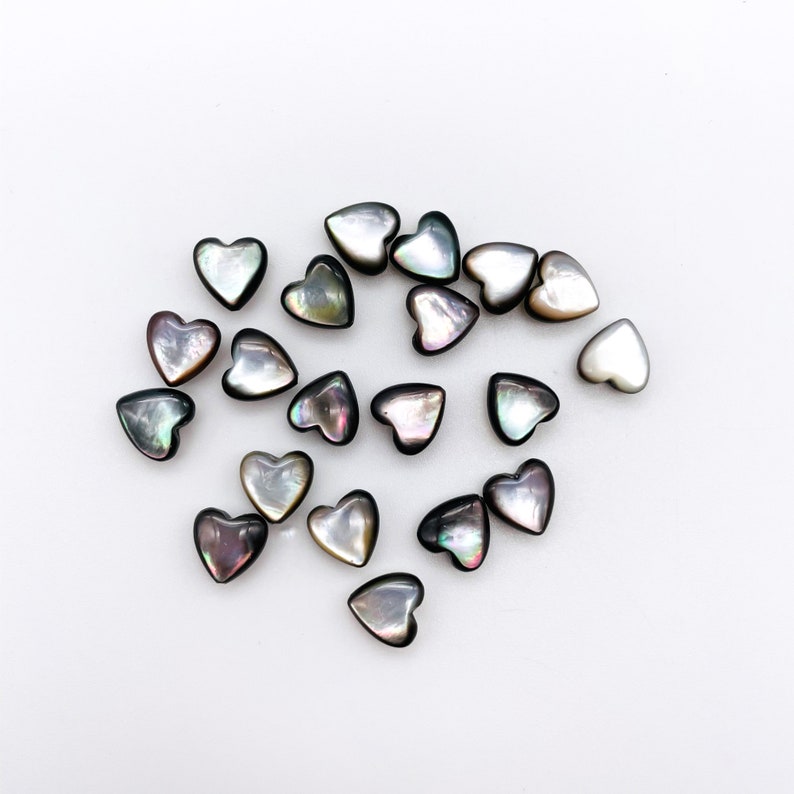 May include: A collection of iridescent black heart-shaped shell beads. The beads are scattered on a white surface.