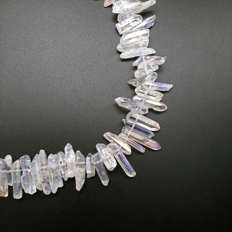 Pode incluir: Uma linha de pontas de cristal branco iridescente, tamb&eacute;m conhecidas como quartzo aura de anjo, dispostas em semic&iacute;rculo sobre um fundo preto.