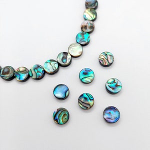 May include: A strand of iridescent abalone shell beads in a necklace style, with several loose beads scattered around. The beads are round and have a blue, green, and gold colour.