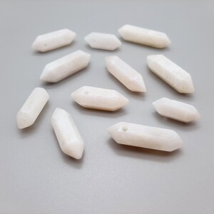 May include: A collection of twelve white, smooth, polished, and elongated gemstone beads with a hole drilled through the center of each bead. The beads are shaped like a pointed oval.