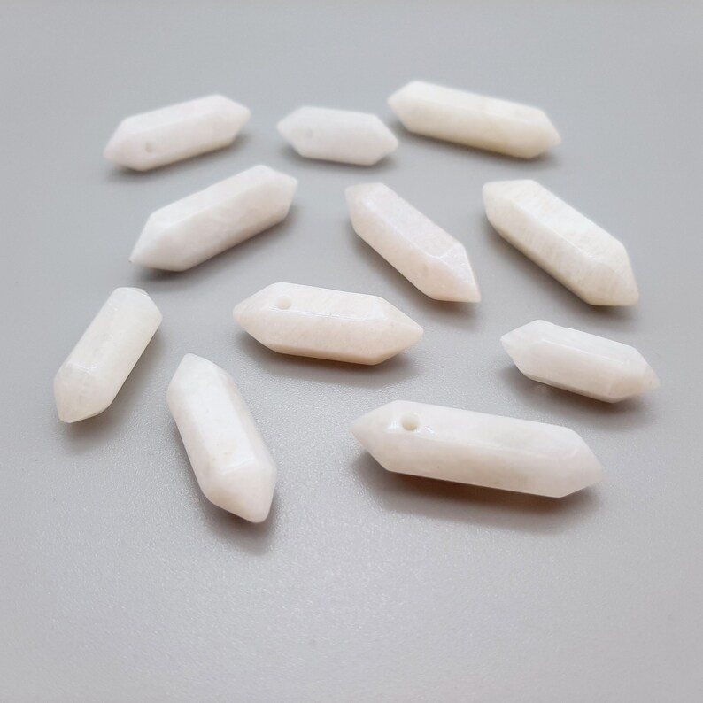 May include: A collection of twelve white, smooth, polished, and elongated gemstone beads with a hole drilled through the center of each bead. The beads are shaped like a pointed oval.