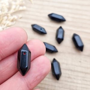 May include: Seven black, faceted, pointed gemstone beads with a hole drilled through the centre. The beads are arranged on a wooden surface.