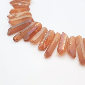 May include: A strand of orange crystal points with iridescent shimmer. The points are arranged in a semi-circle on a white background.