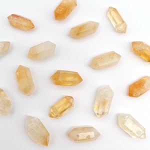 May include: A selection of citrine crystals, in shades of yellow and orange, are arranged on a white background. The crystals are double-terminated, with pointed ends. The crystals are translucent, allowing light to pass through.