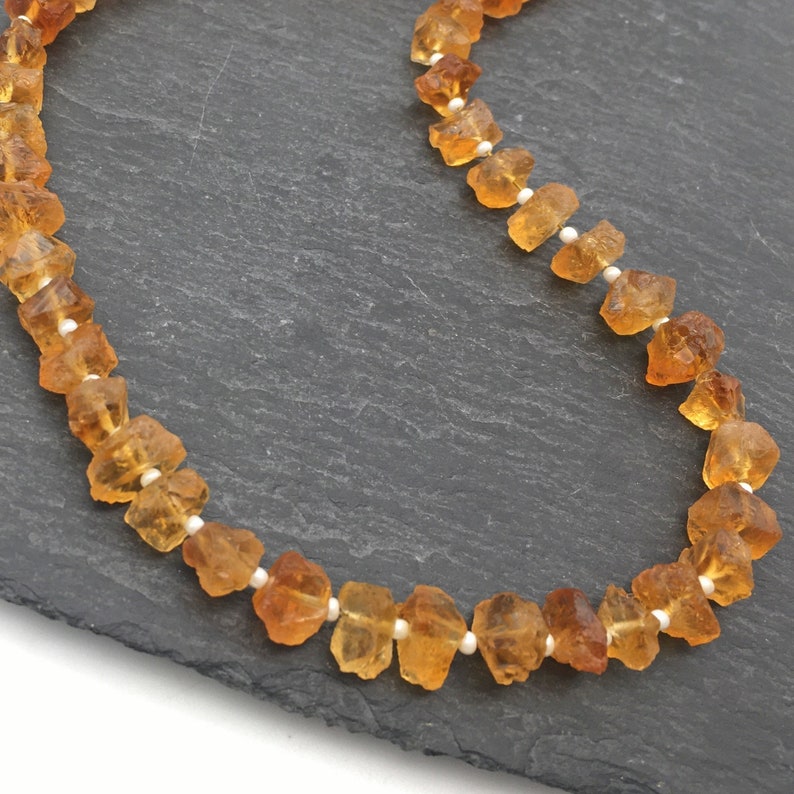 Pu&ograve; includere: Una collana fatta di perline di pietra preziosa arancione e piccole perline bianche. La collana &egrave; su uno sfondo grigio scuro.