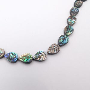 Könnte beinhalten: Eine Kette aus irisierenden, blattförmigen Perlen. Die Perlen haben einen perlmuttartigen Glanz mit Farben von Blau, Grün und Gold. Die Blätter sind aufgereiht und bilden ein dekoratives Element für Schmuck oder Bastelarbeiten.