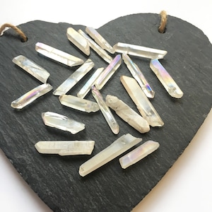 May include: A collection of iridescent white quartz points, also known as angel aura quartz, on a black slate heart-shaped surface.