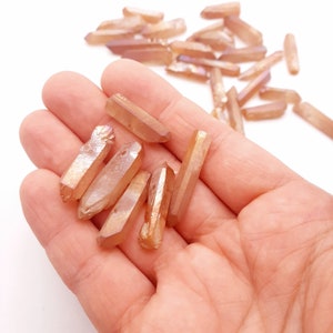 May include: A collection of small, iridescent, orange-brown quartz points. The points are arranged in a hand, with some scattered on a white surface.