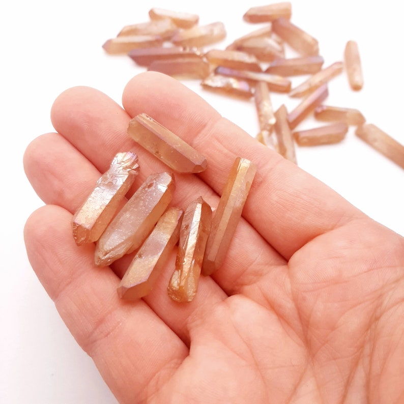 May include: A collection of small, iridescent, orange-brown quartz points. The points are arranged in a hand, with some scattered on a white surface.