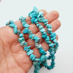 May include: A necklace or bracelet made of turquoise chip beads. The beads are a vibrant turquoise colour with some variations in shade. The beads are irregularly shaped and strung together. The piece is held in a hand against a white background.