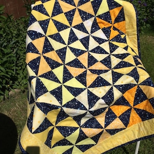 May include: A yellow and orange quilt with a navy blue background. The quilt is made of fabric with a star pattern and a pinwheel design. The quilt is folded and sitting on a white chair.