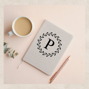 May include: A beige notebook with a black floral wreath and the letter "P" in the center. A cup of coffee and a pen are on a pink surface.