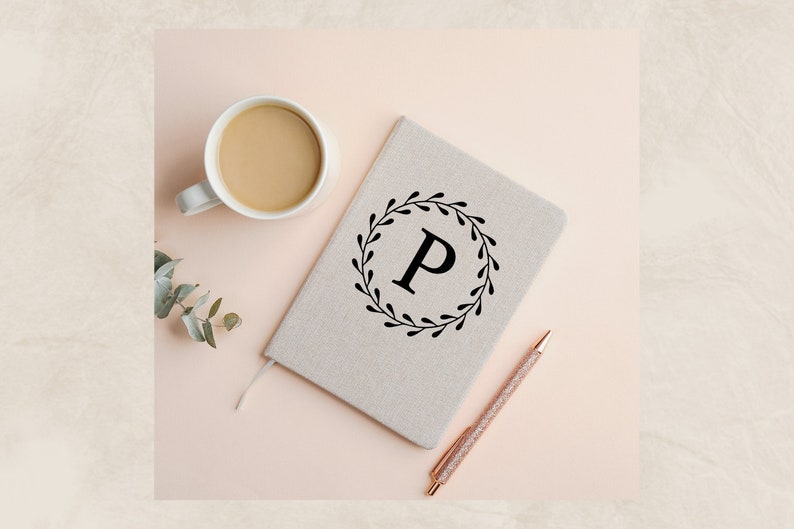 May include: A beige notebook with a black floral wreath and the letter "P" in the center. A cup of coffee and a pen are on a pink surface.