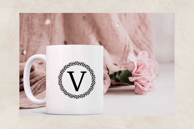 May include: White ceramic mug with a black wreath design and the letter "V" in the center. The mug is sitting on a white surface with a pink fabric and a pink carnation flower.