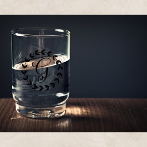 May include: A clear glass with a black and white monogram design featuring the letter "G" inside a wreath of leaves. The glass is filled with water and is sitting on a wooden surface.