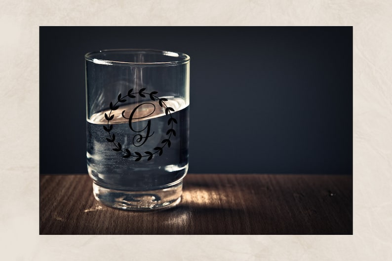 May include: A clear glass with a black and white monogram design featuring the letter "G" inside a wreath of leaves. The glass is filled with water and is sitting on a wooden surface.