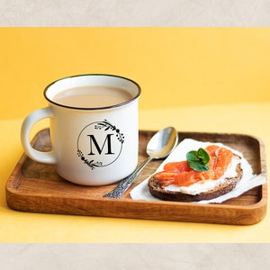May include: A white mug with a black floral design and the letter "M" in a circle on the side, filled with coffee, on a wooden tray with a slice of bread topped with cream cheese and smoked salmon.