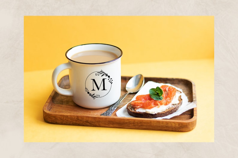 May include: A white mug with a black floral design and the letter "M" in a circle on the side, filled with coffee, on a wooden tray with a slice of bread topped with cream cheese and smoked salmon.