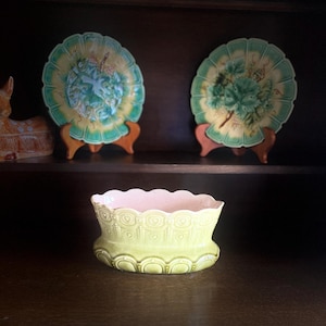 Könnte beinhalten: Zwei dekorative Teller mit gewellten Rändern und floralen Designs, die auf Holzständern ausgestellt sind. Darunter befindet sich ein hellgrüner und rosa Keramiktopf mit gewelltem Rand. Die Teller haben ein gelb-grünes Farbschema.