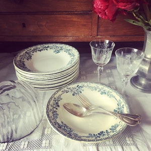 May include: A stack of vintage white ceramic soup bowls with a blue and green floral pattern. The bowls are on a white tablecloth with a stack of plates and a glass cloche.