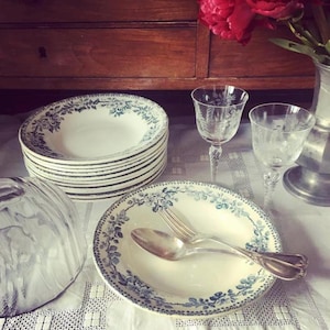 May include: A stack of white ceramic bowls with blue floral patterns, alongside a bowl with a fork and spoon. Two etched wine glasses and a glass dome are also present. The setting is on a white lace tablecloth.