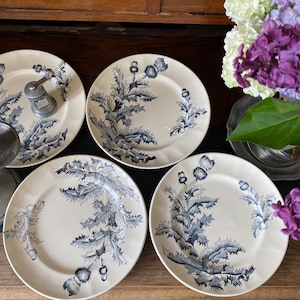 Peut inclure: Ensemble de quatre assiettes en céramique blanche avec un motif de chardon bleu. Les assiettes présentent une esthétique vintage classique, le motif floral couvrant la majeure partie de la surface. Elles sont disposées sur une surface en bois.