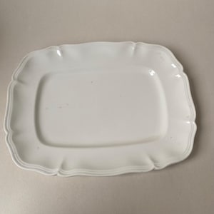 Puede incluir: Una bandeja de servir rectangular de cerámica blanca con esquinas redondeadas y un borde festoneado en relieve. La superficie de la bandeja es lisa y brillante, con un borde ligeramente elevado que enmarca la zona central plana del plato.