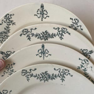 Könnte beinhalten: Ein Stapel antiker weißer Keramikteller mit einem tealfarbenen Blumen- und Banddesign. Die Teller weisen ein sich wiederholendes Muster aus Blumen, Bändern und einem fackelförmigen Emblem auf. Die Teller haben eine Vintage-Ästhetik.