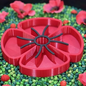 May include: A red poppy-shaped mould for making cookies or other treats. The mould is made of plastic and has a detailed design. It is sitting on a bed of green and red sprinkles.