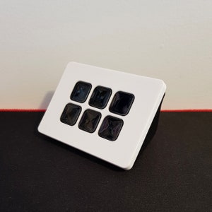 以下が含まれることがあります： 6つの黒いボタンが付いた白い長方形のデバイス。ボタンは2行3列に配置されています。デバイスは右側に少し傾いています。