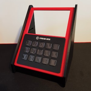 May include: A red and black Stream Deck stand with a black Stream Deck device. The stand has a raised back and a flat front. The Stream Deck has 15 buttons with a black background and clear plastic covers.