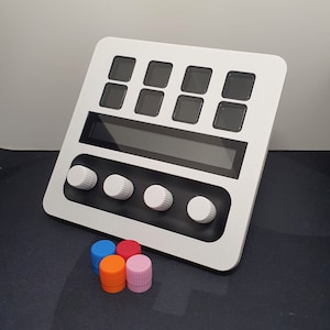 May include: White electronic device with a black screen and eight square buttons. There are four white knobs on the bottom of the device. Four colourful plastic caps are in front of the device.