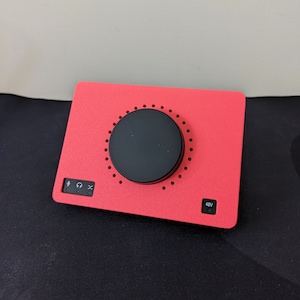 May include: A red audio interface with a large black knob in the centre. The interface has a microphone input, headphone output, and a 48V button.