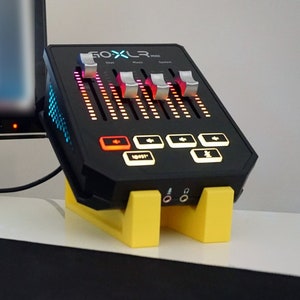 Op de afbeelding: Een zwarte GOXLR Mini audiomixer met vijf faders, elk met een bijbehorende LED-lichtstrip. De mixer heeft een gele standaard en verschillende knoppen met LED-verlichting. De tekst "GOXLR MINI" staat op de voorkant van de mixer.