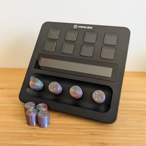 May include: A black Stream Deck device with a screen and eight buttons. The device has four round knobs with iridescent rainbow coloured tops. There are five additional iridescent rainbow coloured knobs on a wooden surface.