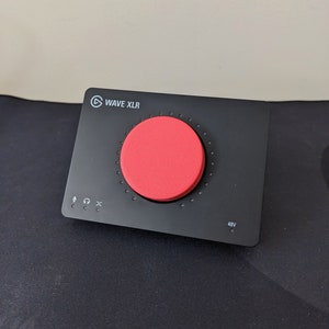 Puede incluir: Una interfaz de audio negra Wave XLR con una perilla roja y un botón de 48V. La interfaz tiene una entrada de micrófono, un conector para auriculares y un botón de silencio.