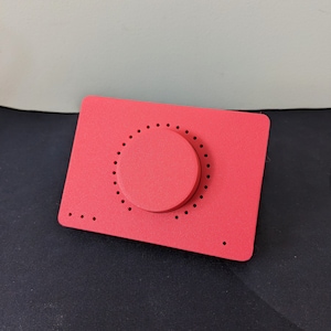 Könnte beinhalten: Ein rechteckiges rotes Gerät mit einem großen runden Knopf in der Mitte. Der Knopf hat ein kreisförmiges Muster aus kleinen Punkten.