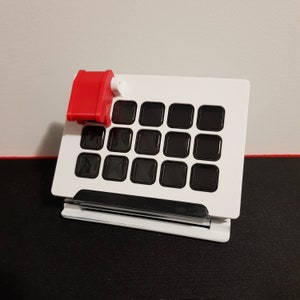 Könnte beinhalten: Eine weiße und schwarze Tastatur mit einem roten Kippschalter. Die Tastatur hat ein Raster aus 16 schwarzen Quadraten.