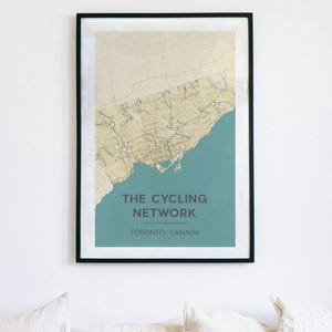 May include: Framed print of a map of Toronto's cycling network. The map is in beige and blue, with the text "THE CYCLING NETWORK" and "TORONTO, CANADA" in a dark font. The frame is black.