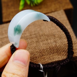 May include: A jadeite crescent moon charm bracelet with a dark brown braided cord. The jadeite is a light green colour with white and dark green swirls.