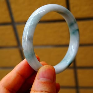 May include: A jade bangle bracelet with a smooth, polished surface. The bracelet is mainly white with streaks of pale green and a translucent quality. The circular design is held by a hand, showcasing the bracelet's shape and colour variations.
