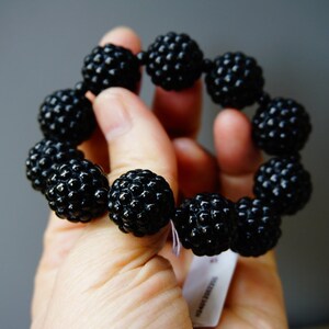 May include: A black beaded bracelet with a textured surface. The bracelet is made of multiple round beads strung together. The beads have a bumpy, almost berry-like appearance. The bracelet is held in a hand against a gray background.