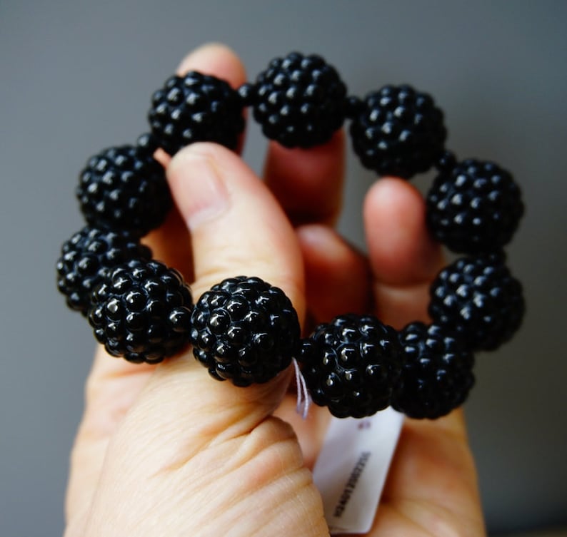 May include: A black beaded bracelet with a textured surface. The bracelet is made of multiple round beads strung together. The beads have a bumpy, almost berry-like appearance. The bracelet is held in a hand against a gray background.