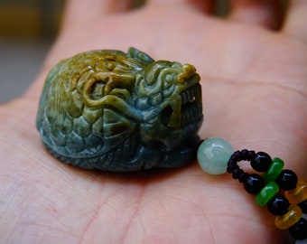 May include: A jade green dragon pendant with brown and yellow accents, carved in detail. The pendant is attached to a black and green beaded cord.