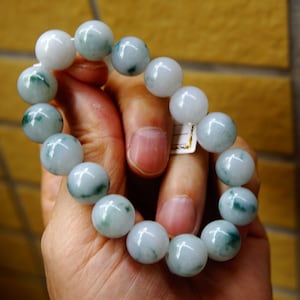 May include: A bracelet made of large, round beads in white and green hues. The beads have a marbled effect, with darker green swirls and patches. The bracelet is held in a hand, highlighting the smooth, polished texture of the stones.
