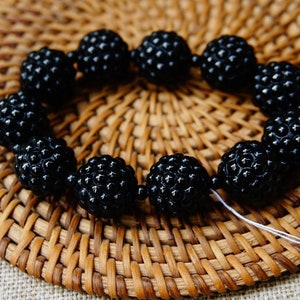 May include: A bracelet made of black, textured beads, arranged in a circular shape. The beads have a bumpy surface and are strung together with a white string. The bracelet is set against a woven, brown background.